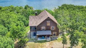 A two-story wooden house with a large porch and a covered outdoor seating area, surrounded by dense green trees and a body of water in the background.