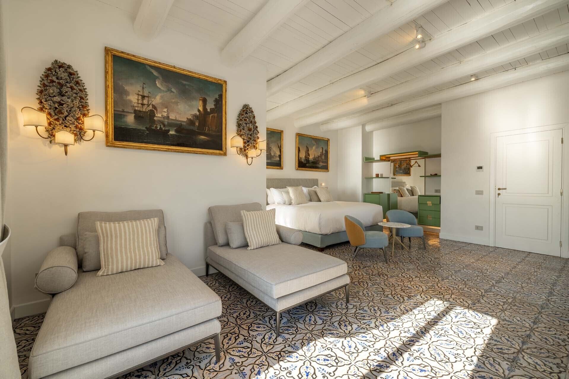 Une chambre d’hôtel avec un lit, deux canapés, une petite table et une chaise. On y trouve des lampes fixées au mur et des tableaux.