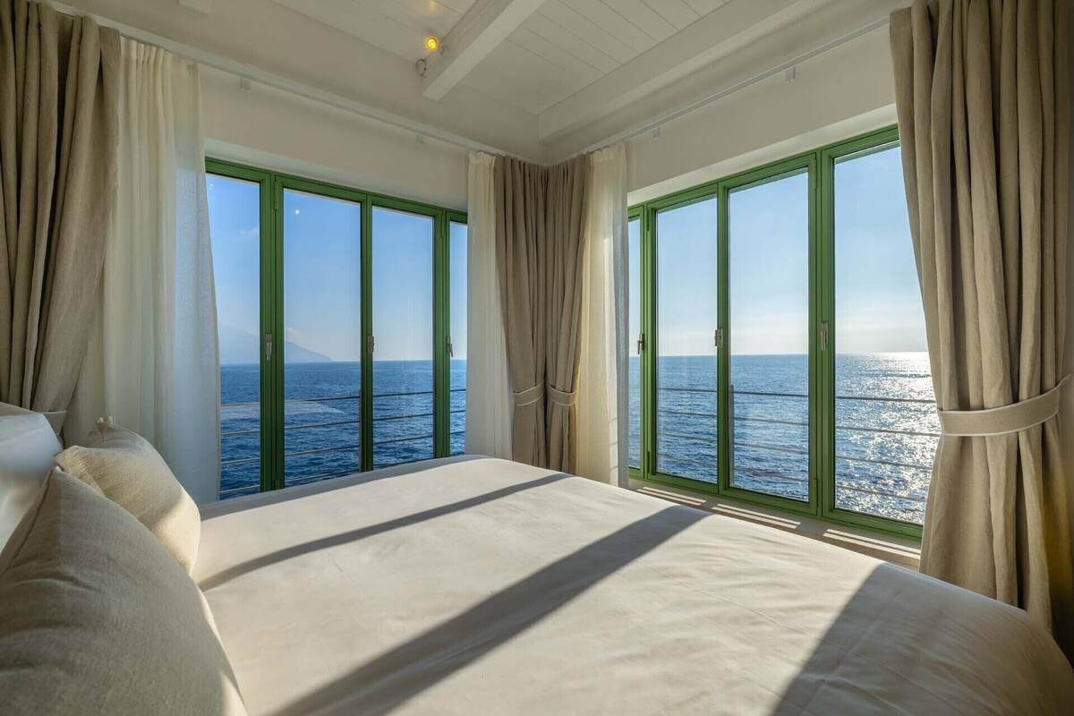 A bedroom with a large bed, two windows with curtains, and a view of the sea.