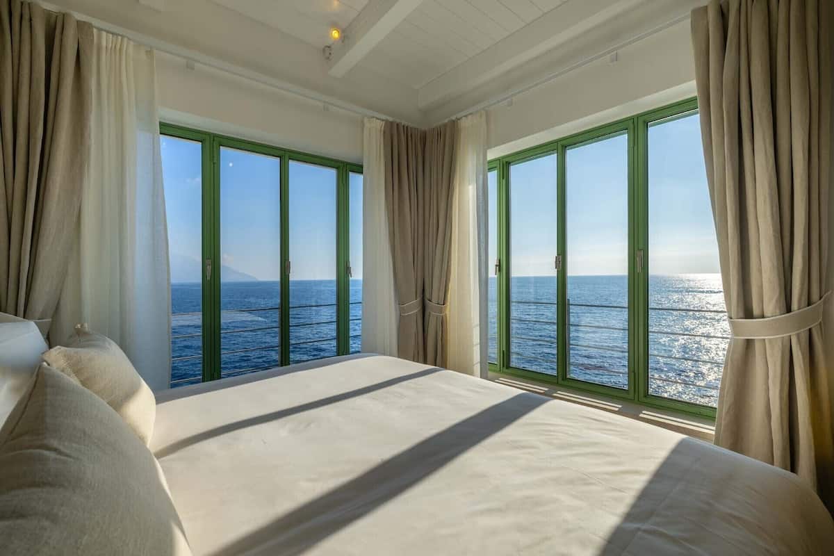 A bedroom with a large bed, two windows with curtains, and a view of the sea.