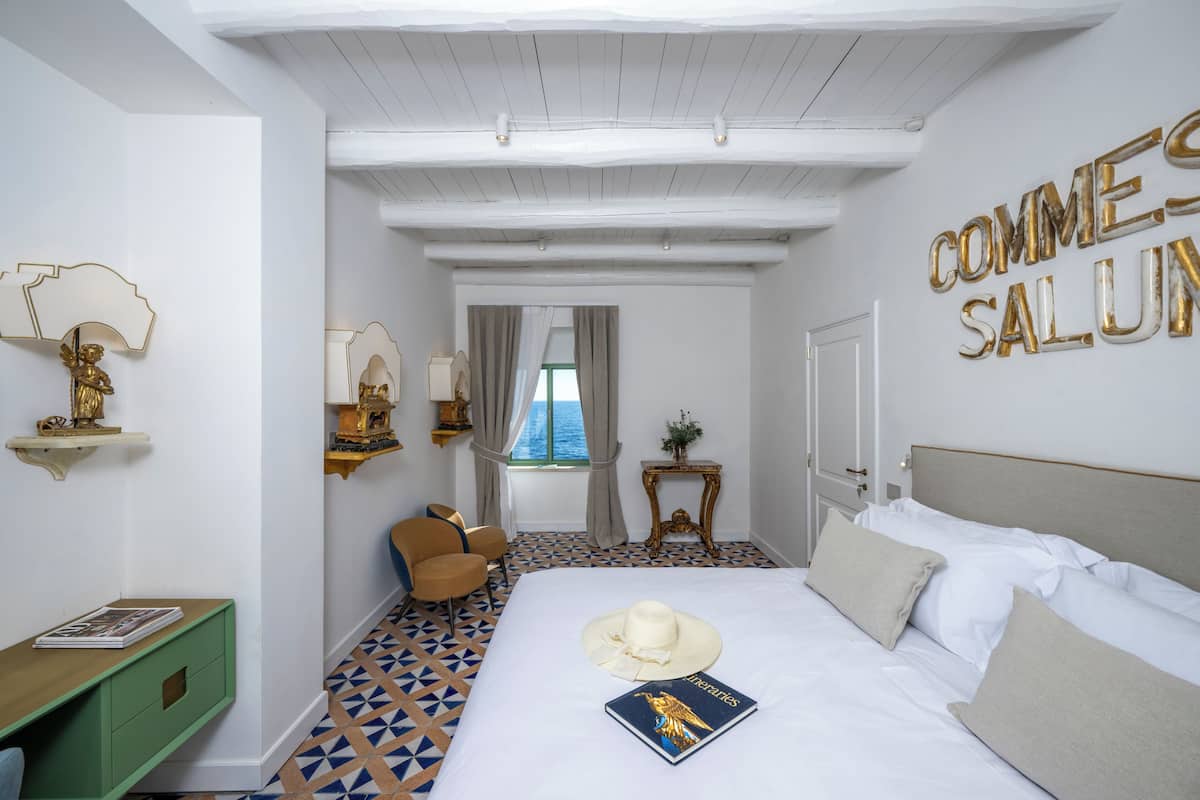 A hotel room with a bed, a chair, a small table, and a view of the sea through a window.