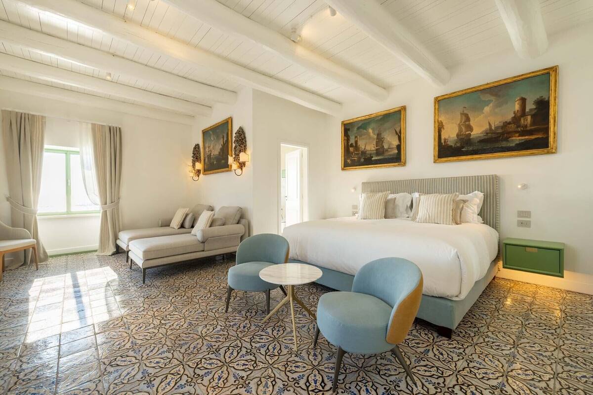 A hotel room with a bed, a sofa, a chair, and two paintings on the wall.