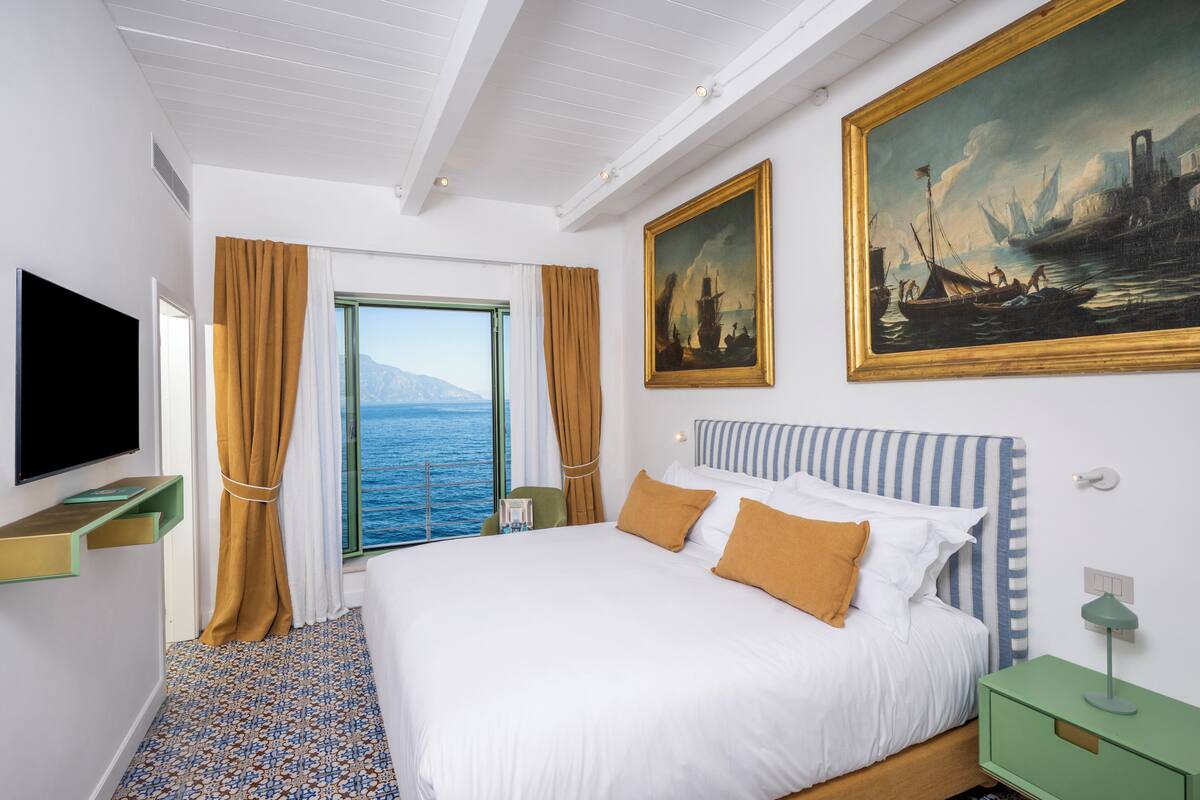 A hotel room with a bed, a TV, a painting, and a view of the sea.