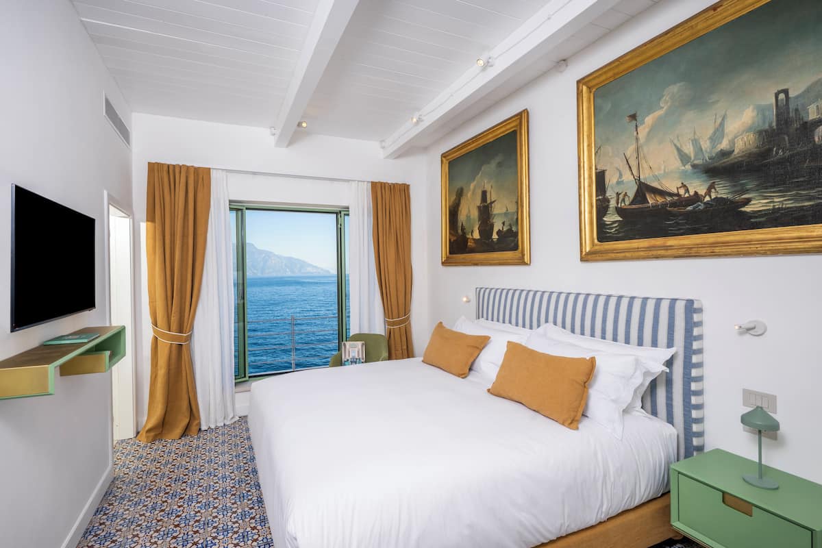 A hotel room with a bed, a TV, a painting, and a view of the sea.