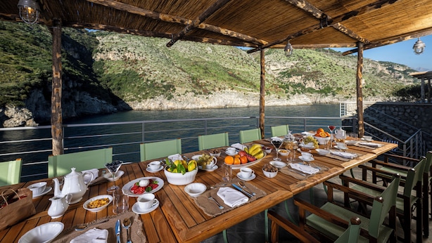 Una mesa de madera preparada para el desayuno, con vistas al mar y las colinas.