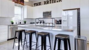 Une cuisine moderne avec des meubles blancs, un îlot central doté de tabourets, un éclairage suspendu et un réfrigérateur en acier inoxydable.