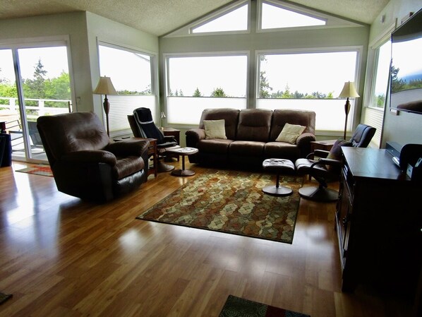A spacious living room with a brown leather sofa, a brown armchair, and a coffee table. There are two lamps, a rug, and a TV on a stand.
