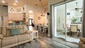 A spacious living room with a beige sofa, a dining area with a table and chairs, and a kitchen visible through a glass door. - "Shore Fun, Bare Feet Retreat" (Spring Hill)