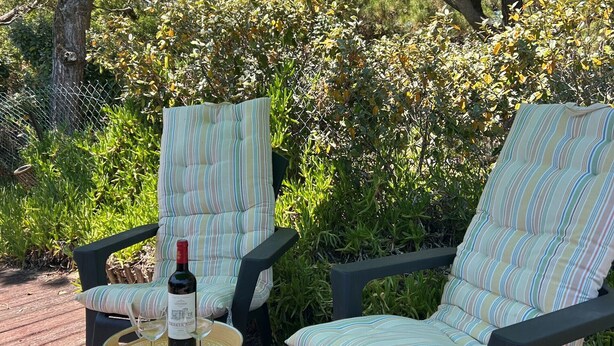 Dos sillas a rayas bajo un paraguas verde sobre una terraza de madera, con una botella de vino en una mesa redonda.