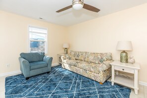 Une pièce dotée d’un ventilateur de plafond, d’un canapé bleu à motifs, d’un fauteuil bleu clair, d’un tapis à motifs et d’une petite table de chevet blanche avec une lampe.