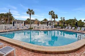 A swimming pool with lounge chairs and umbrellas, surrounded by palm trees and buildings.