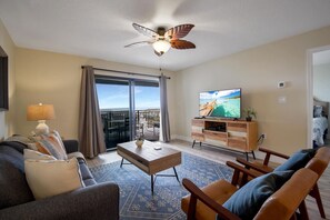 A living room with a sofa, coffee table, and TV, a balcony with a view, and a ceiling fan.