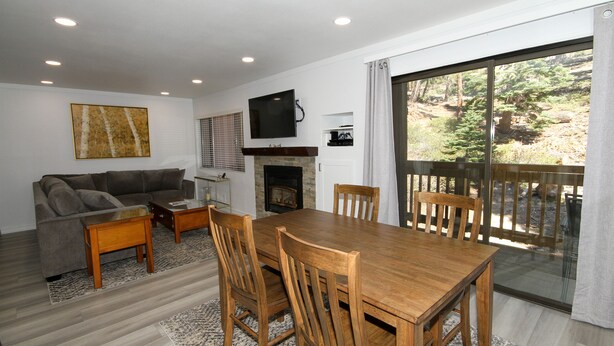 A living room with a sofa, coffee table, wooden dining table, and chairs. There's a fireplace, a TV mounted on the wall, and a large window with a view of trees. - Mammoth Ski & Racquet Club #127 (Mammoth Lakes)