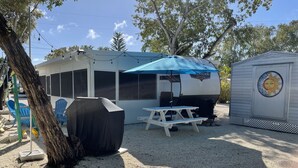 A covered RV with a picnic table and a grill, a covered barbecue area, and a small shed with a decorative sun emblem. - The place to go when you want to get away! (Key Largo)