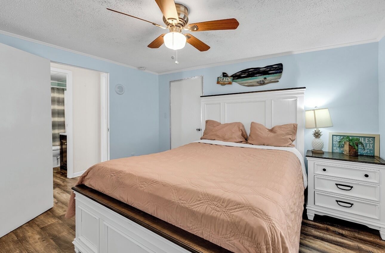 A bedroom with a bed, a nightstand, a ceiling fan, a door, and a wall with a decorative item.