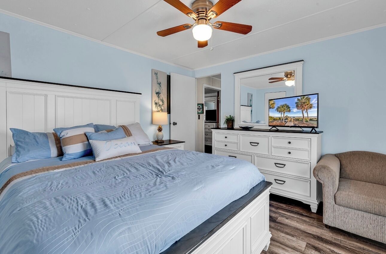 Une chambre à coucher comprenant un lit, une commode, une chaise, un ventilateur de plafond et un téléviseur.