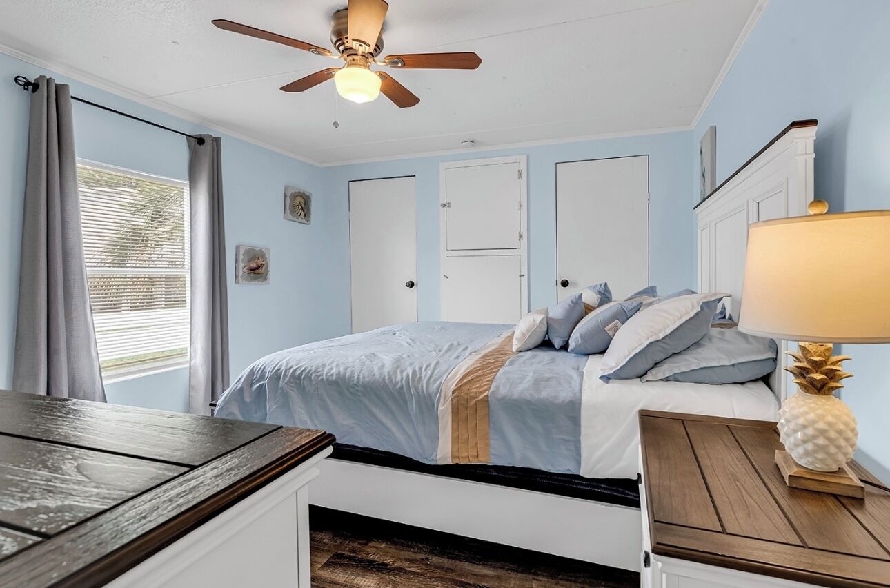 Un dormitorio con cama, ventilador de techo, mesita de noche y ventana con cortinas.