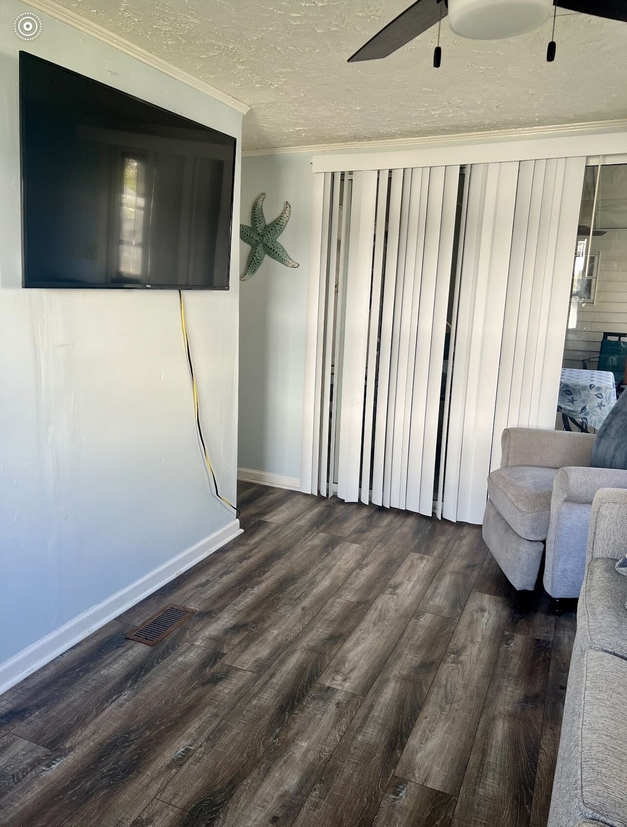 A living room with a flat-screen TV mounted on the wall, a starfish decoration, vertical blinds, a grey sofa, and a wooden floor.