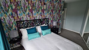 Een slaapkamer met een groot bed, een kleurrijk abstracte muur, een kroonluchter en een nachtkastje met een lamp.