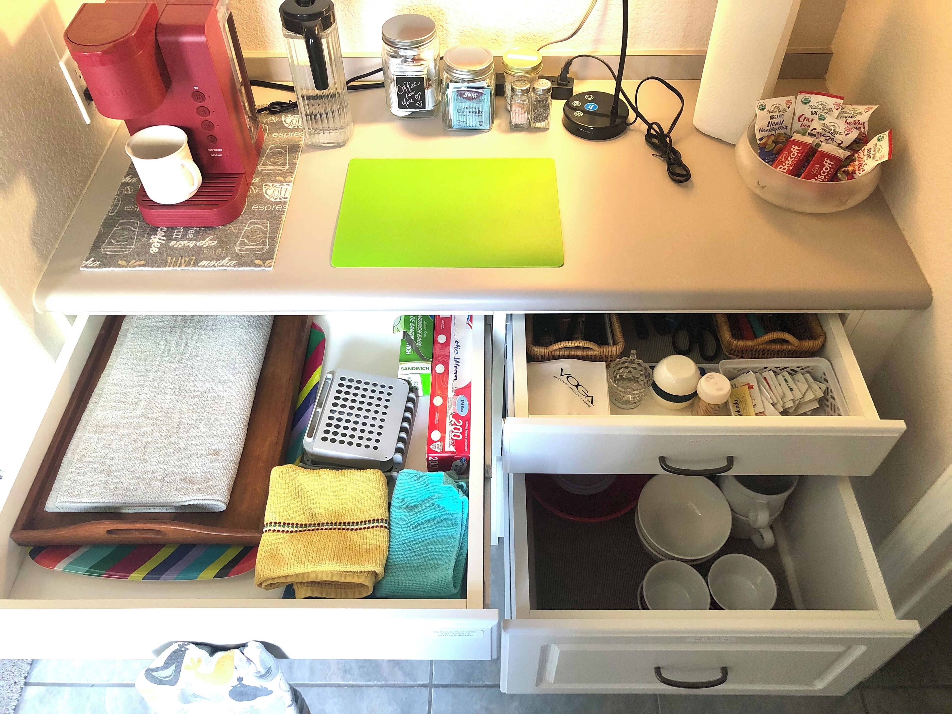 A kitchen counter with a coffee maker, jars, and a bowl of snacks. Below, a drawer contains kitchen utensils, a cutting board, and a towel.