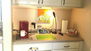 A small kitchenette with white cabinets, a coffee maker, jars, a green cutting board, and a fire extinguisher.