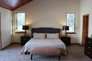 A bedroom with a large bed, two bedside tables with lamps, a bench, and two windows with a view of trees. - Glacier Majesty · Getaway Next Door to Glacier National Park (West Glacier)