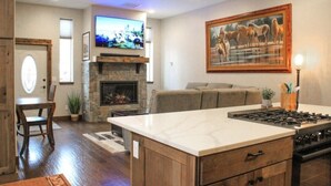 Una cocina moderna con chimenea de piedra, un comedor con mesa y sillas de madera, y una sala de estar con sofá y televisor montado.