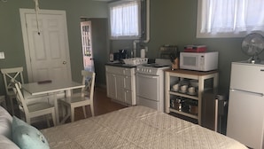 Een compacte kamer met een bed, een kleine kitchenette, een eettafel met stoelen en een deur naar een andere kamer.