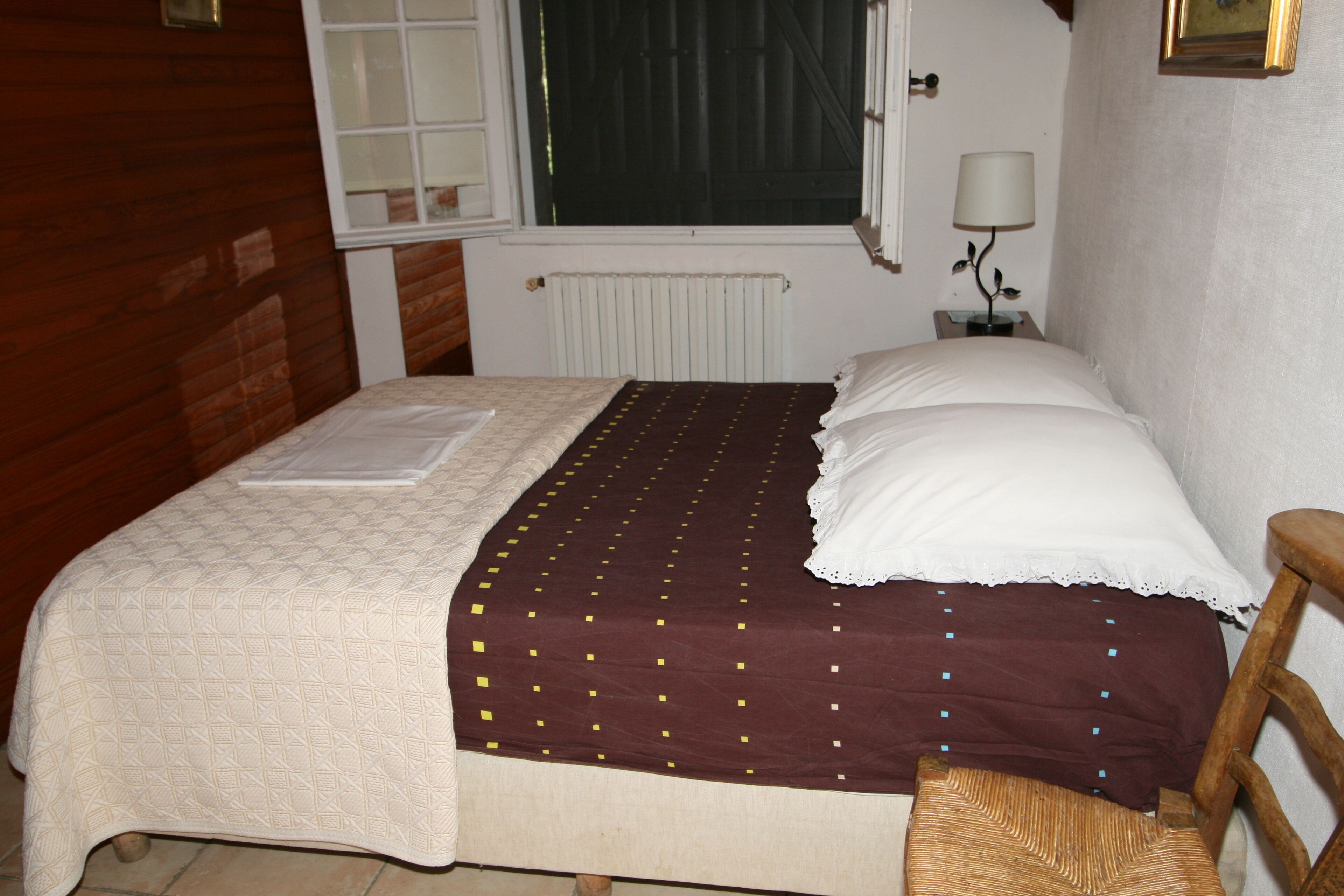 Un lit recouvert d’une couvre-lit à motifs, une table de chevet avec une lampe et une fenêtre donnant sur l’extérieur.