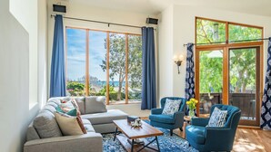 A spacious living room with a grey sofa, a blue armchair, and a wooden coffee table. Large windows offer a view of greenery and a building. - Large Home with Ocean Views & Hot Tub! (Santa Barbara)