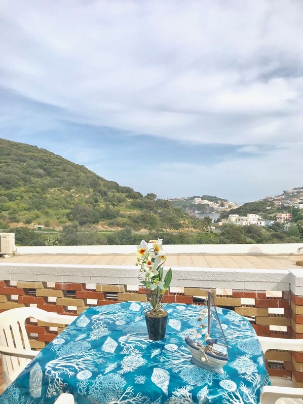 Une table recouverte d’une nappe bleue, une orchidée en pot et un fauteuil suspendu sur une terrasse en toit, d’où l’on peut admirer une ville perchée sur une colline.