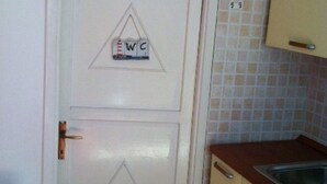 Une porte blanche ornée d’un symbole triangulaire et des lettres « WC » indiquant les toilettes. Une petite table recouverte d’une nappe en dentelle se trouve à gauche de la porte. On aperçoit, à droite, un coin cuisine avec un évier et des placards.