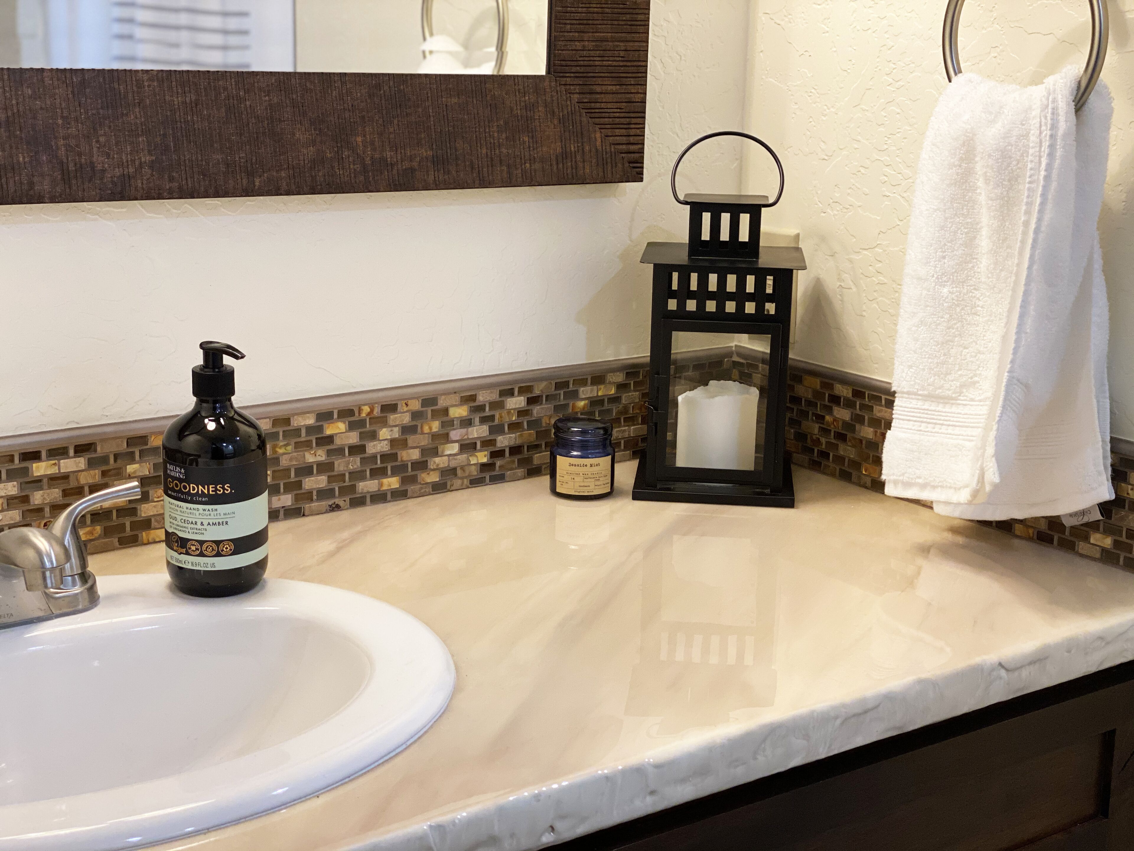 Un lavabo de salle de bain avec un flacon de savon liquide, une petite bougie et une serviette suspendue à un anneau métallique.