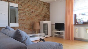 A living room with a brick wall, a grey sofa, a TV on a stand, and a window with curtains. - Country house style meets modernity (Mühlheim am Main)