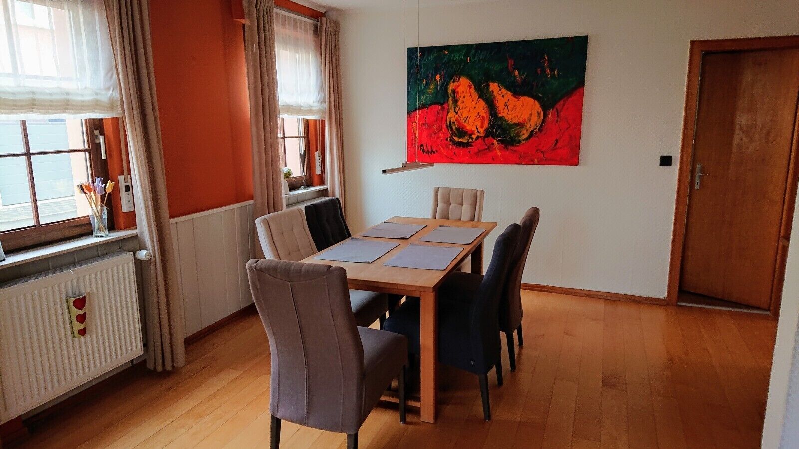 A dining area with a wooden table, six chairs, a radiator, a window with curtains, and a painting on the wall.