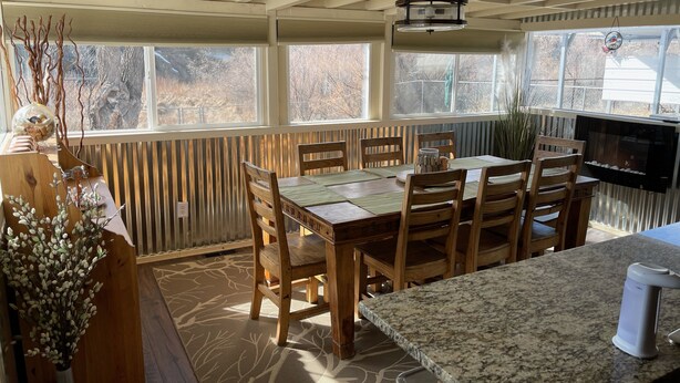 A dining area with a wooden table and chairs, a granite countertop, and a view of the outdoors through large windows.
