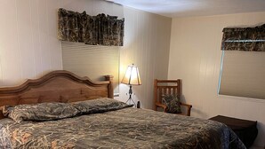 A bedroom with a large bed, a nightstand, a lamp, a chair, and a window with blinds.