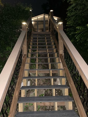 Un escalier en bois, doté de rampes en métal et d’éclairages en haut, entouré de verdure.
