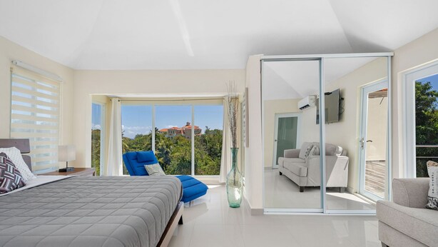 Un dormitorio moderno con una cama grande, una silla azul y una puerta de vidrio que da a un balcón con vistas a zonas verdes y edificios.