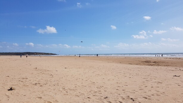 Una amplia playa de arena con huellas y algunas personas a lo lejos.