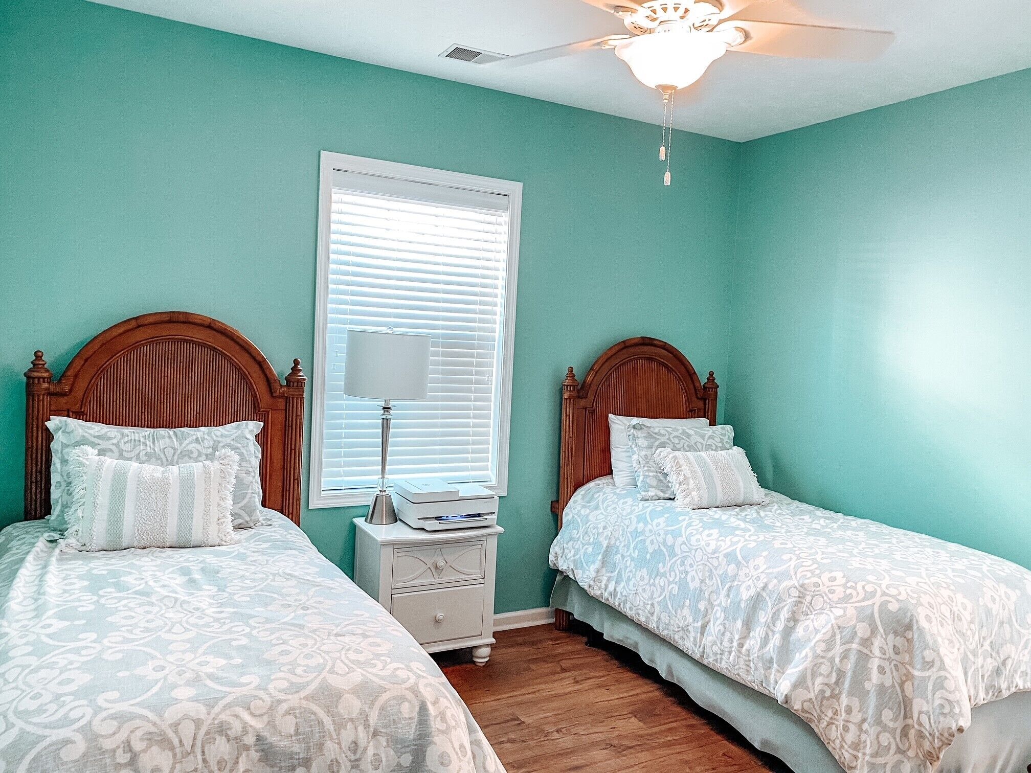 Quarto com duas camas, uma mesinha de cabeceira, um ventilador de teto e uma janela com persianas.