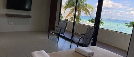 Habitación de hotel con cama, dos sillones, televisor y balcón con vista al océano y palmeras.