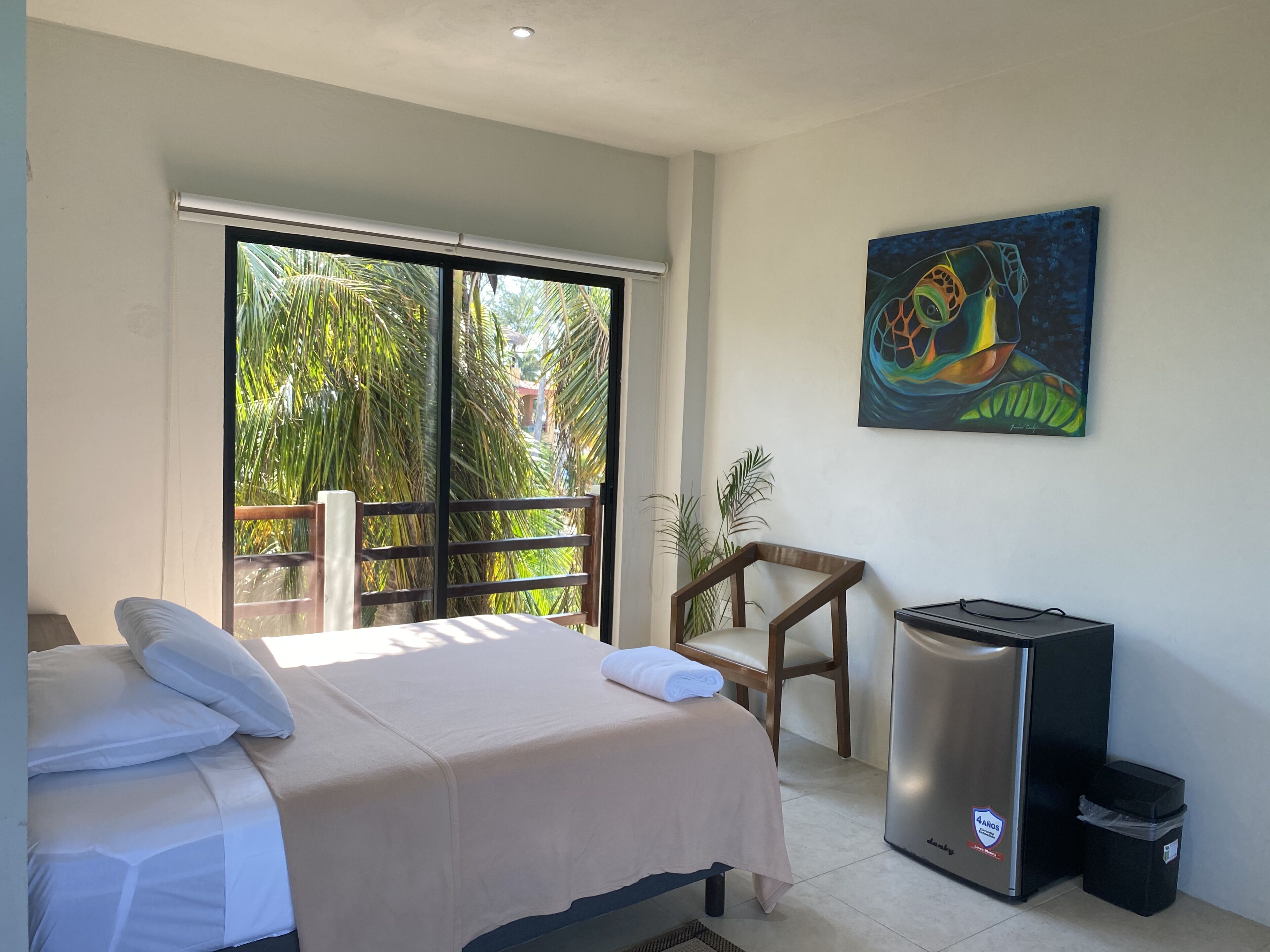 Habitación de hotel con una cama, una silla, un cesto de basura y un cuadro en la pared.
