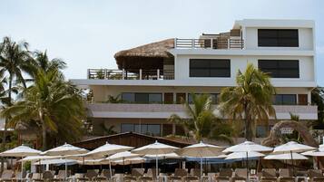 Un hotel frente a la playa con varios pisos, un área de piscina y palmeras.