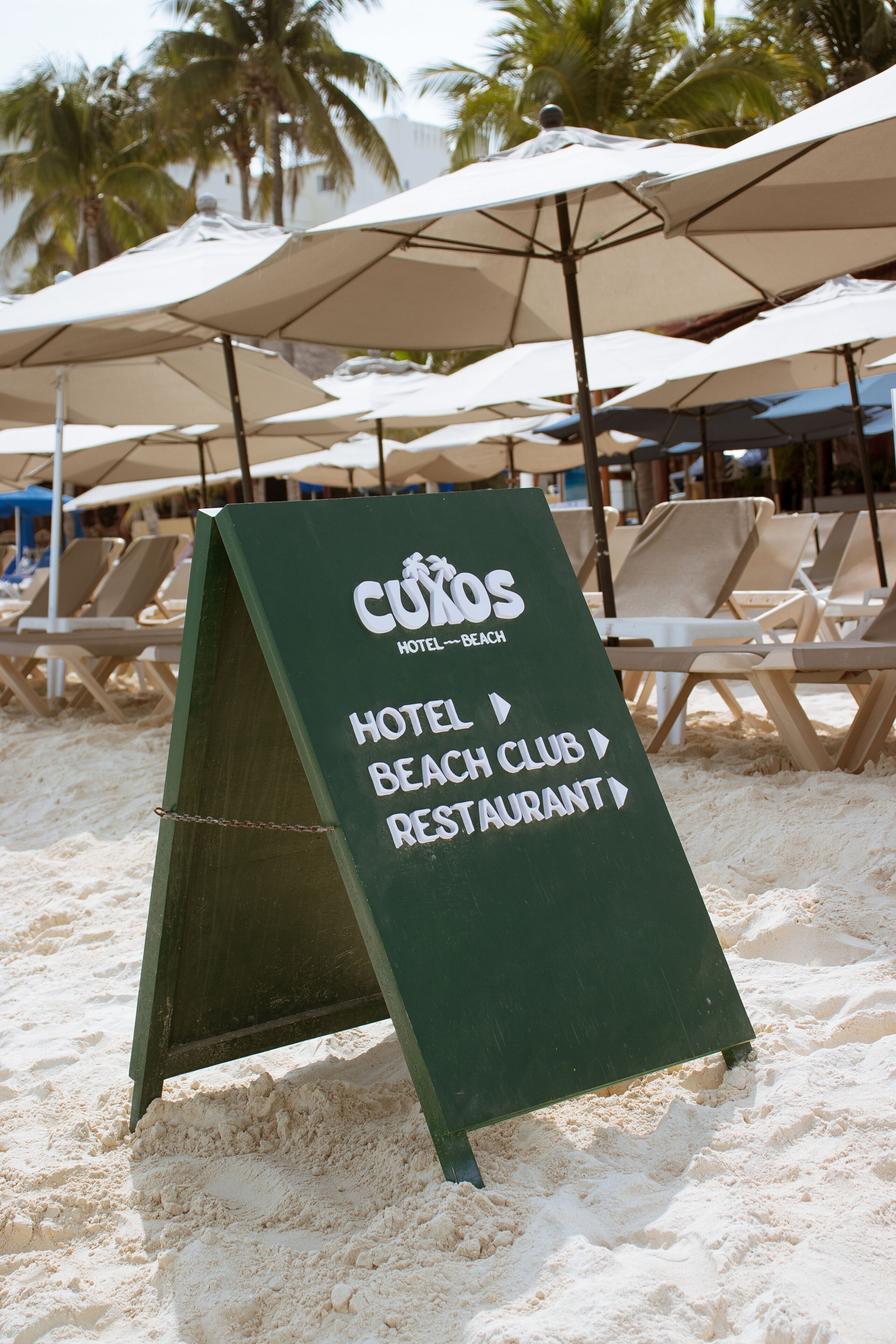 A directional sign on a beach with 'CUXOS HOTEL BEACH CLUB RESTAURANT' written on it.
