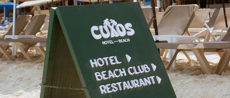 A directional sign on a beach with 'CUXOS HOTEL BEACH CLUB RESTAURANT' written on it.