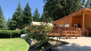 A wooden cabin with a porch, a staircase leading up to it, and a garden area with a small tree and a bench. - Lake Nahwatzel Resort, Cabin #1, Shared Dock just steps away! Now with WIFI! (Shelton)