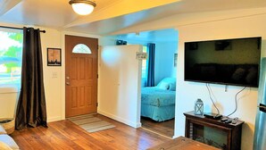 A cozy room with a wooden floor, a bed with a blue bedspread, a television mounted on the wall, and a door leading to another room. - Lake Nahwatzel Resort, Cabin #1, Shared Dock just steps away! Now with WIFI! (Shelton)