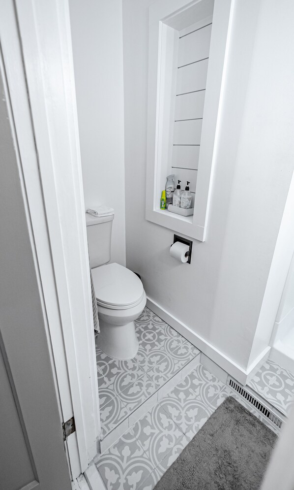 Un baño pequeño y blanco con inodoro, dispensador de papel higiénico y estante con artículos de aseo.