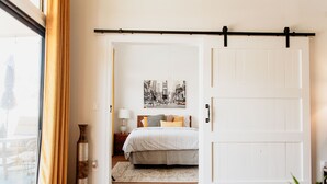 Une chambre à coucher comprenant un lit, une table de chevet, une lampe, une grande fenêtre avec des rideaux et une porte.